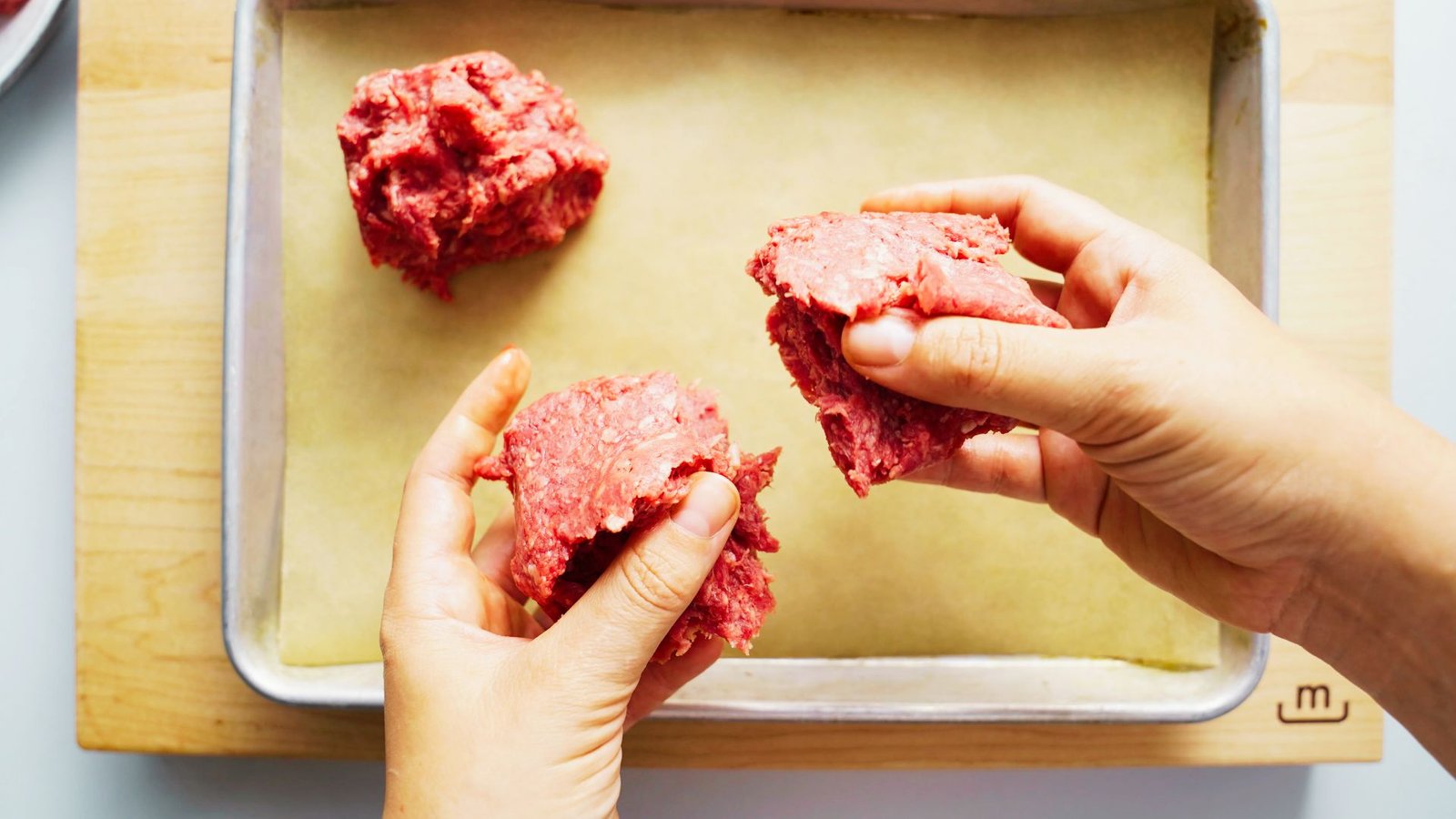 A hand grips a piece of ground beef on a baking sheet, illustrating the process of preparing burger patties.