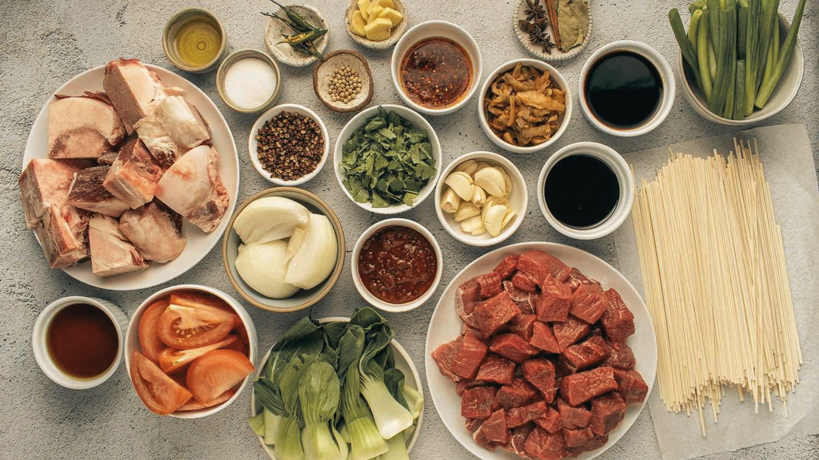 Ingredients for beef noodle soup: a variety of meats, colorful vegetables, and noodles displayed together on a table.