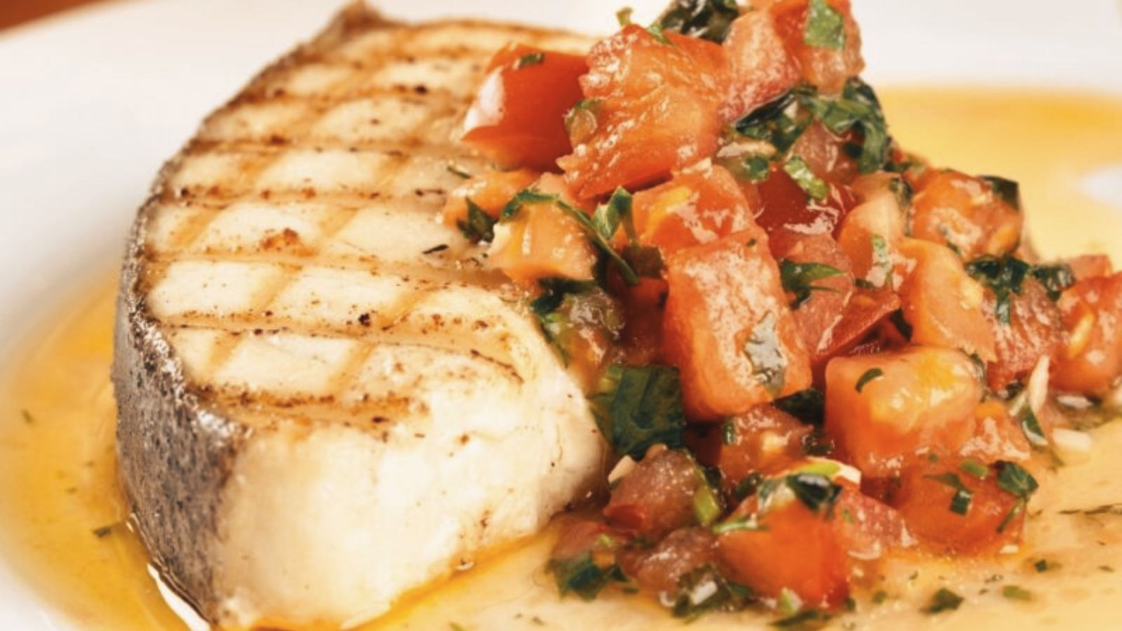 A white plate features grilled fish smothered in vibrant tomato sauce, emphasizing a fresh and tangy taste.