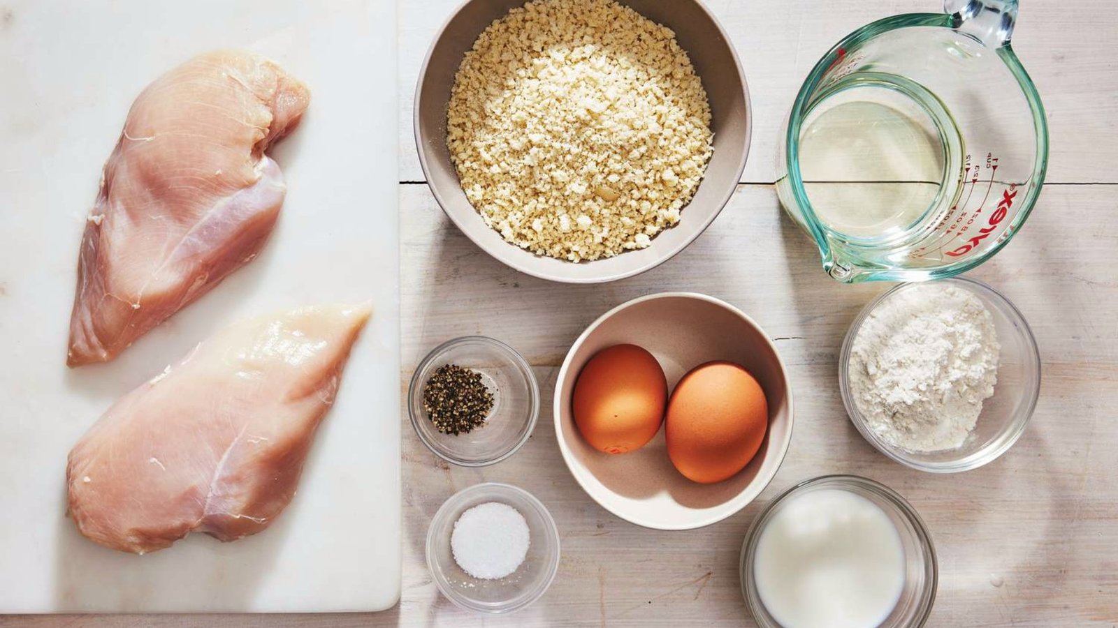 Ingredients for chicken cutlets displayed on a cutting board, including chicken breast, spices, and vegetables.