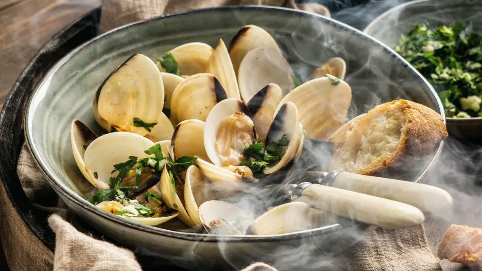 Steamed little neck clams in a bowl, accompanied by fresh bread and herbs, showcasing a delicious seafood dish.