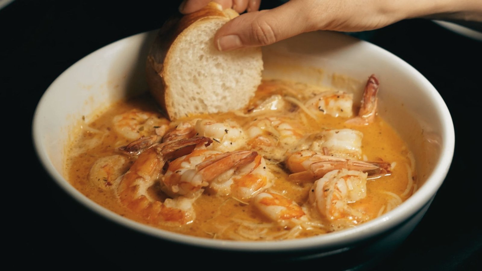 A person holds a piece of bread above a bowl of shrimp, highlighting the flavors of the famous Killer Shrimp recipe.