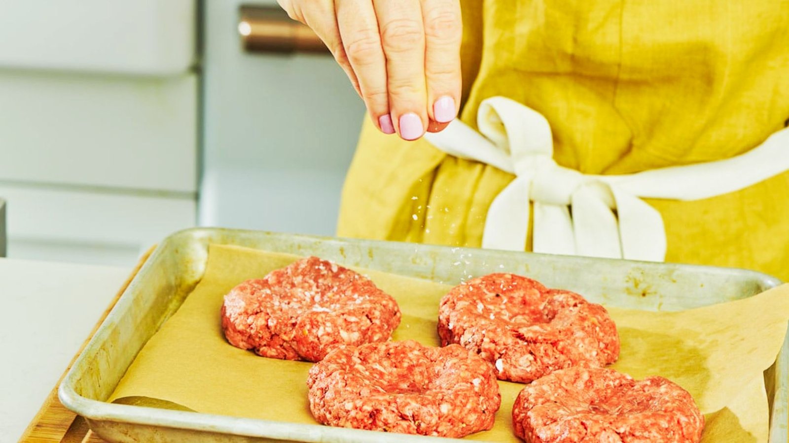 A woman sprinkles ingredients on ground beef while preparing to shape it for burgers.