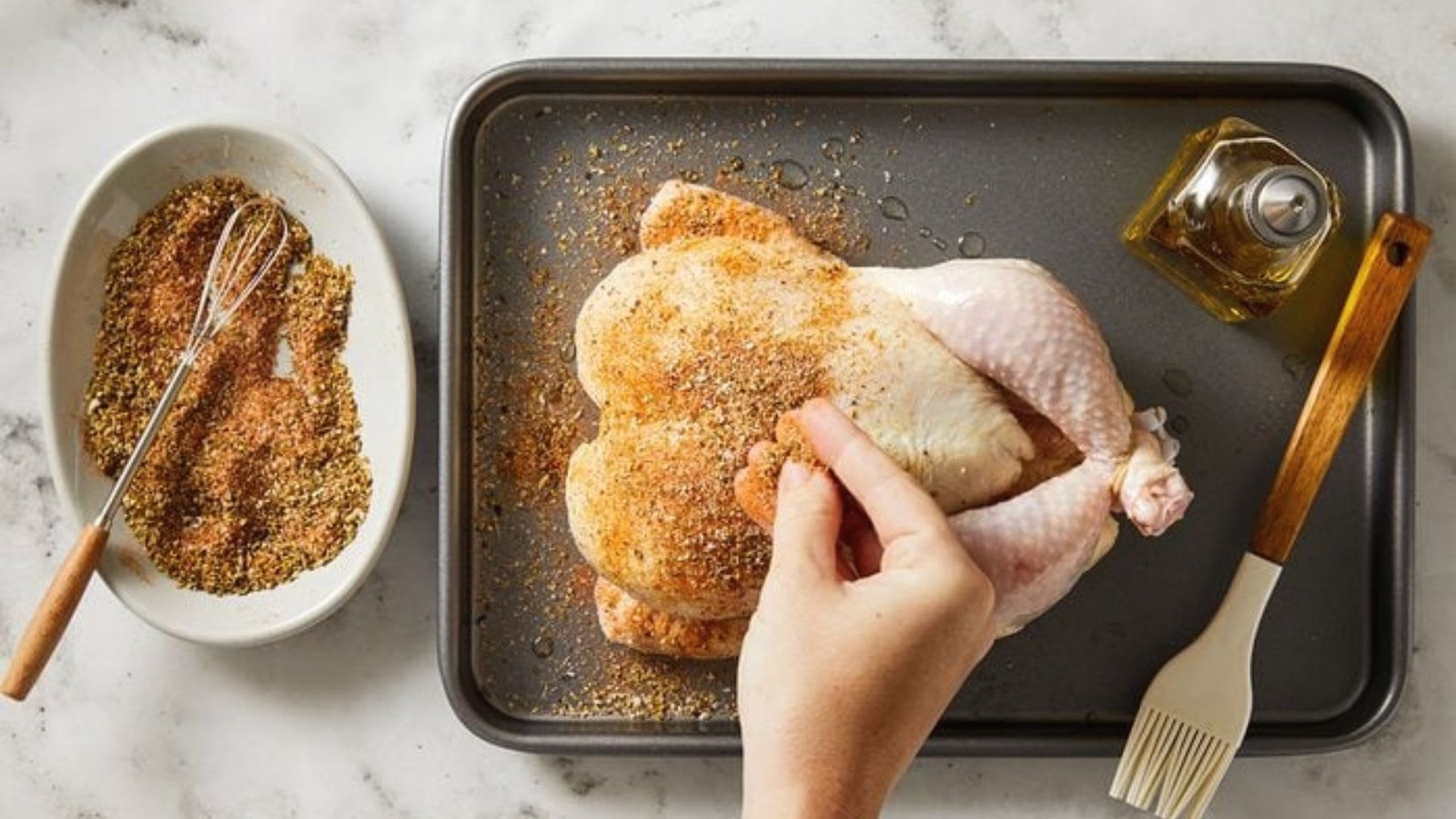 A person is holding a chicken on a baking sheet, indicating the initial step of preparing and seasoning for cooking.