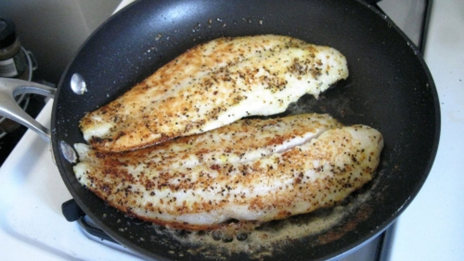Two swai fish fillets cooking in a frying pan on a stove, seasoned with lemon and garlic butter.