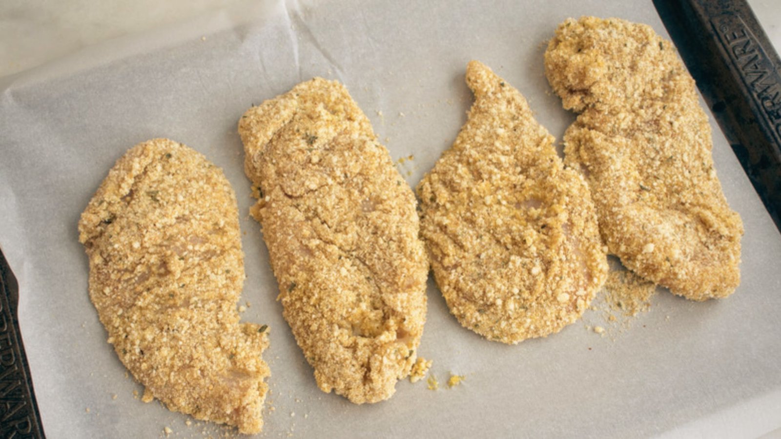 A baking sheet holds four pieces of breaded chicken, set to rest before the cooking process begins.