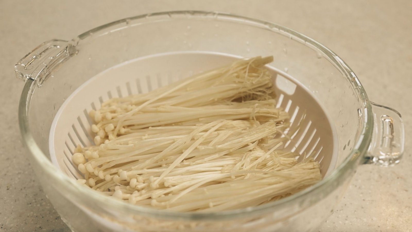 A bowl filled with Enoki Mushrooms resting in a strainer, part of the process for cleaning and preparing enoki mushrooms.