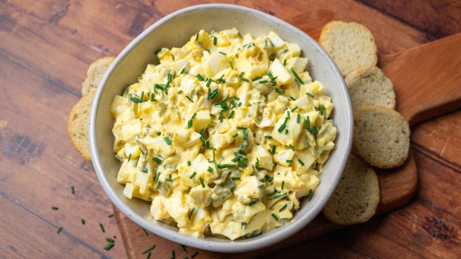 Egg salad in a bowl on a wooden cutting board, garnished with pickles, ready for serving.