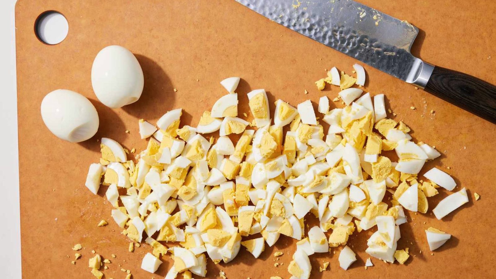 Chopped eggs on a wooden cutting board beside a knife, ready for preparation.