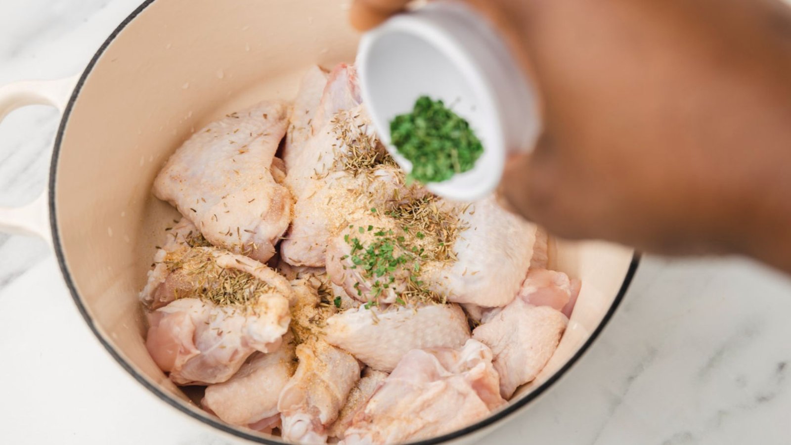 A person is seasoning chicken in a pot by sprinkling herbs, preparing the boiling base for cooking.