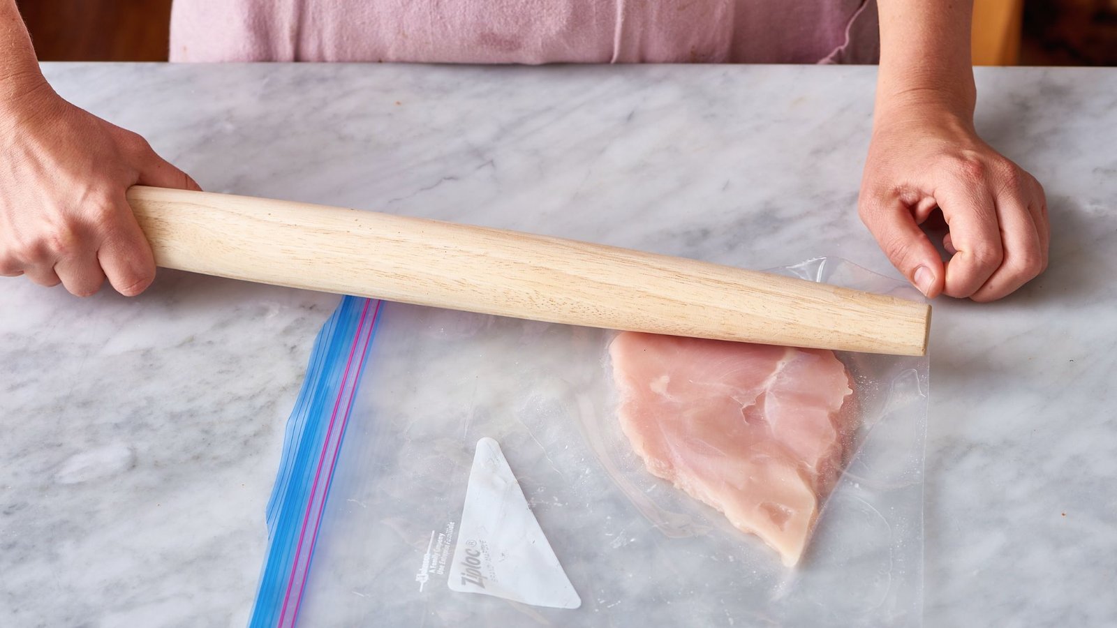 A person rolling up a thinly pounded piece of chicken on a kitchen counter, preparing it for cooking.