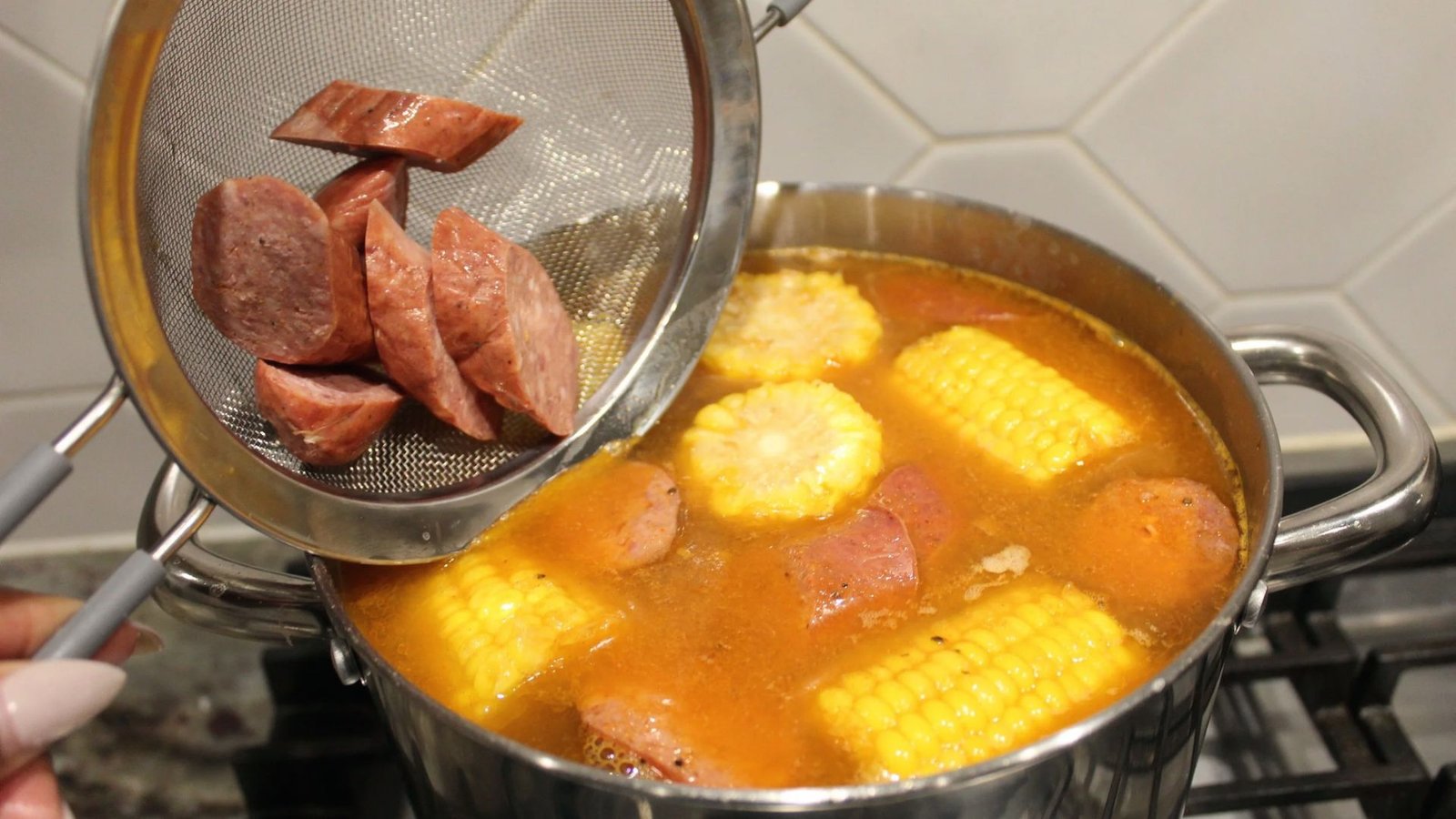 A person stirs corn and sausage into a pot, preparing ingredients for a chicken wing boil recipe.