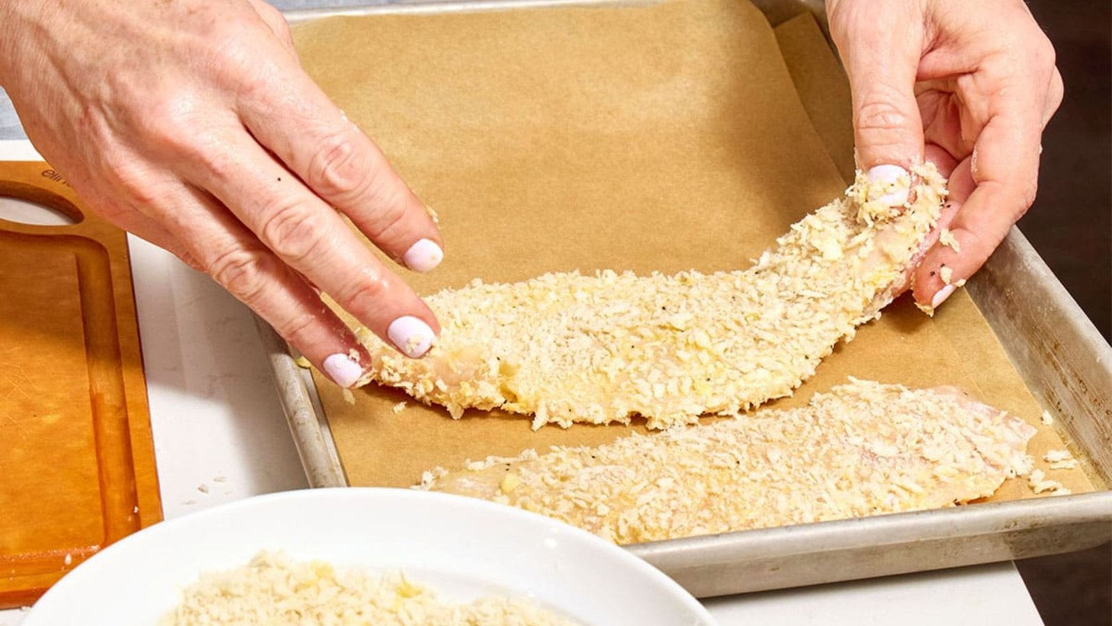 A hand is shown placing a breaded chicken fillet onto a baking plate, illustrating the step for achieving a crispy crust.