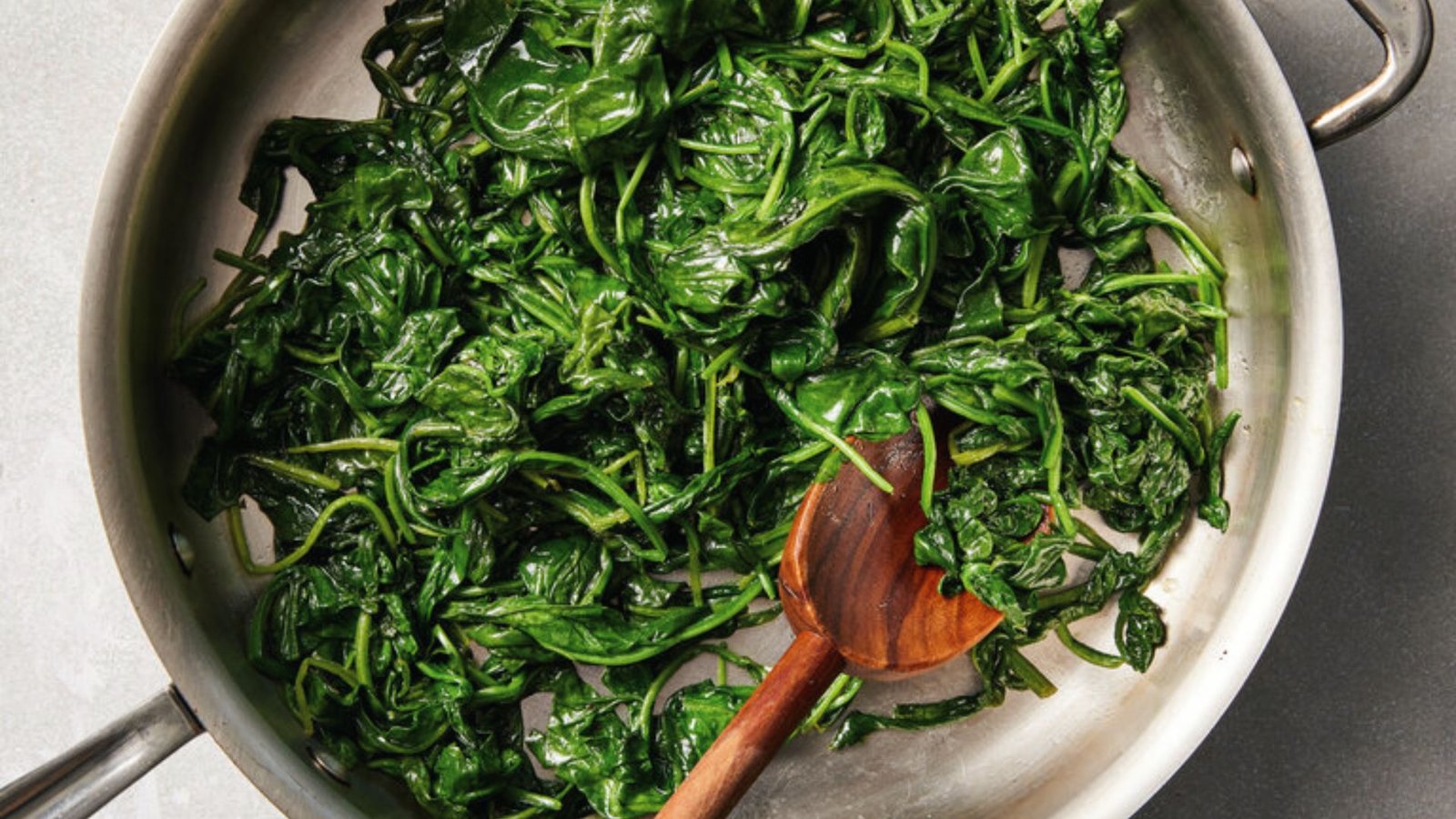 Fresh spinach being sautéed in a pan with a wooden spoon, part of a spinach and shallot mixture preparation.