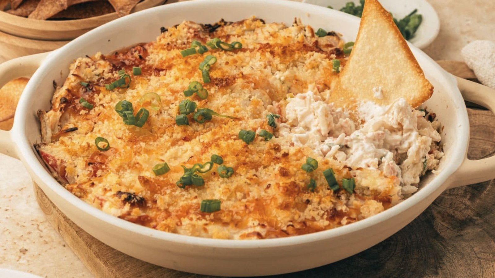 A bowl of hot baked crab rangoon dip surrounded by chips and crackers on a wooden table.