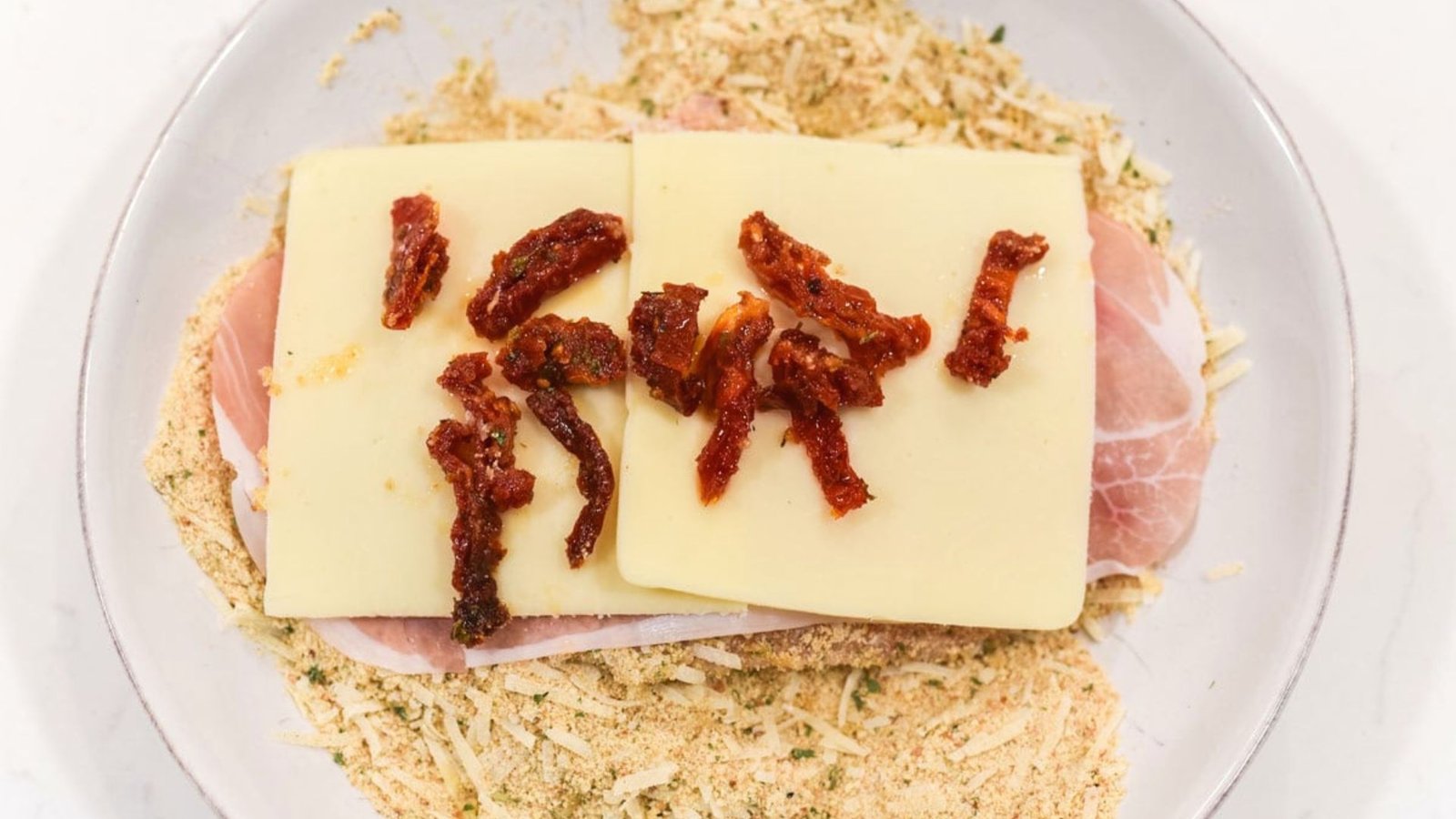 In a white plate full of ingredients that is chicken, cheese, prosciutto, breadcrumb.