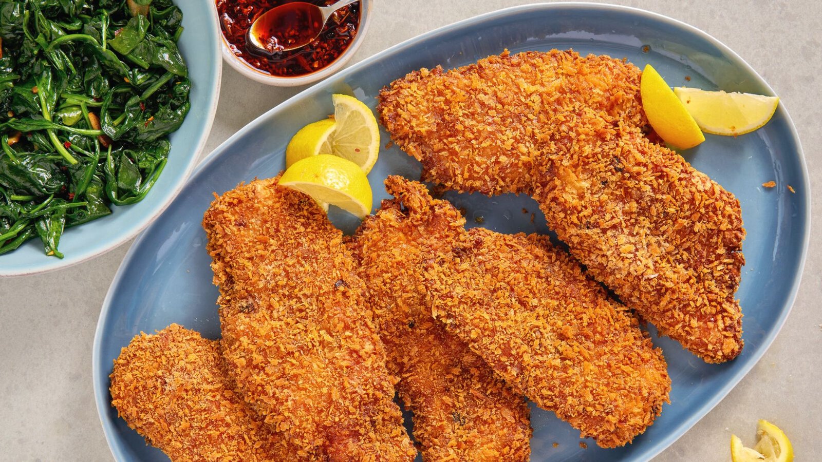 Fried chicken cutlets accompanied by lemon wedges and a side of sautéed spinach on a white plate.