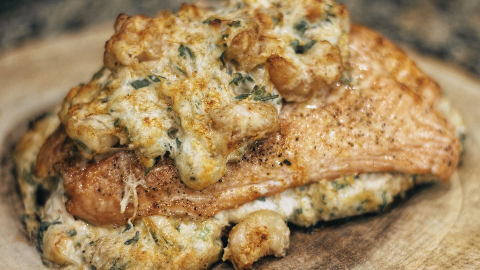 A piece of stuffed salmon topped with cheese and herbs, displayed on a wooden board, showcasing a seafood recipe.