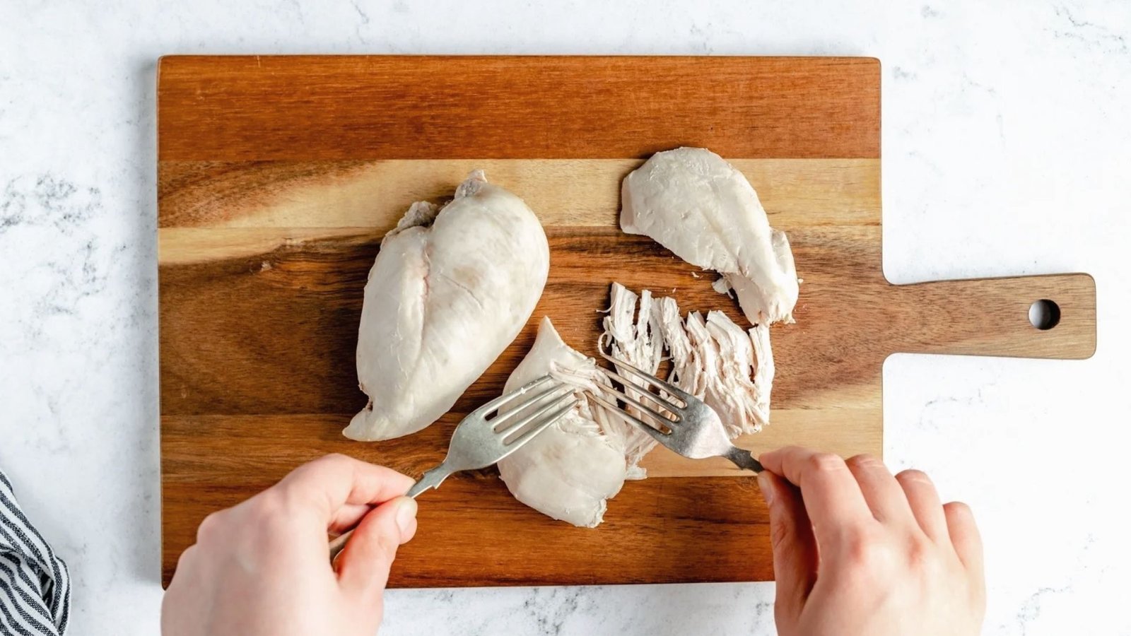 A person slicing chicken on a wooden cutting board, preparing it for shredding.