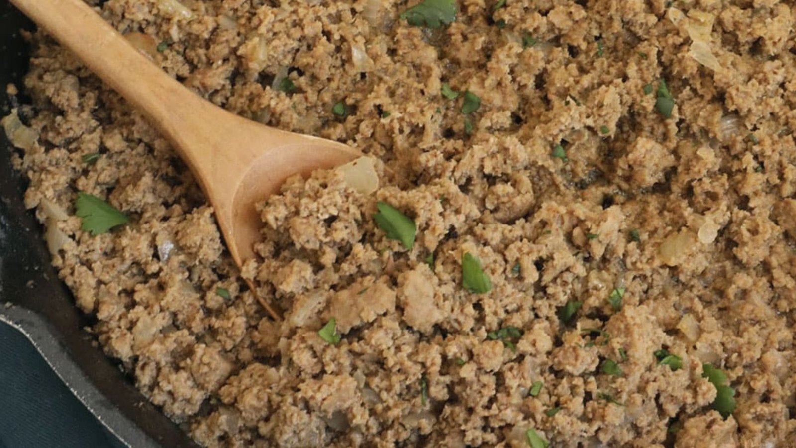 A pan filled with browning ground turkey, accompanied by a wooden spoon for stirring.