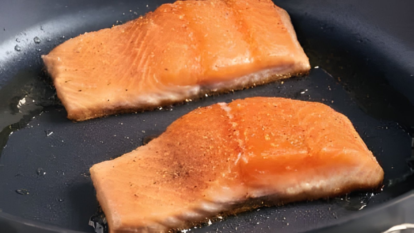 Two pieces of salmon searing in a frying pan over high heat, showcasing a golden-brown crust forming.