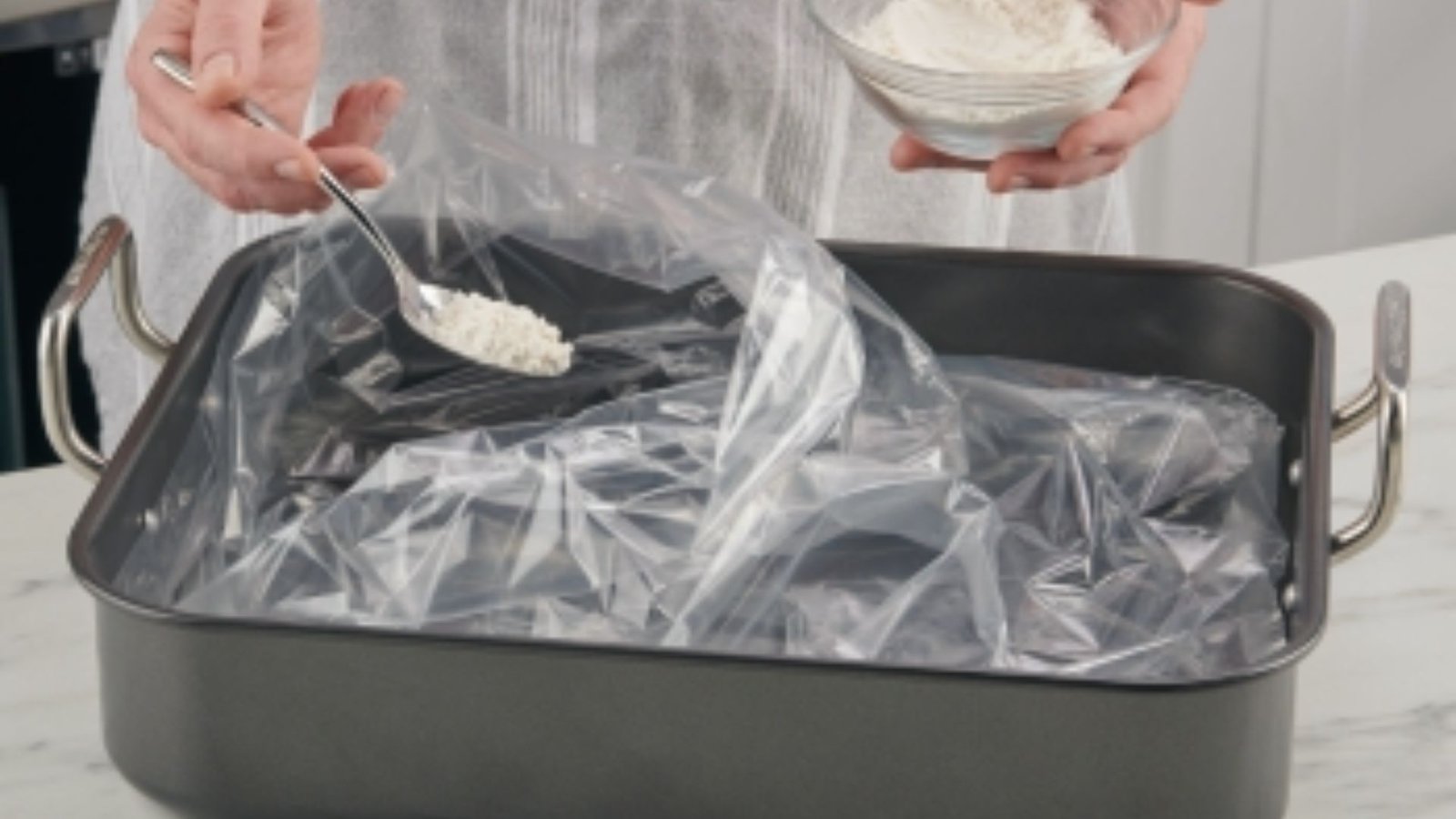 A person is placing a bowl of flour into a plastic bag to prepare an oven bag for cooking.